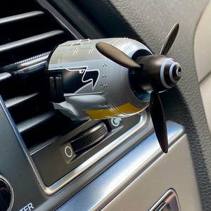 May include: A car air vent clip-on featuring a miniature airplane propeller design. The propeller is black, with a gray and yellow engine casing. The air vent clip is installed in a car's dashboard.