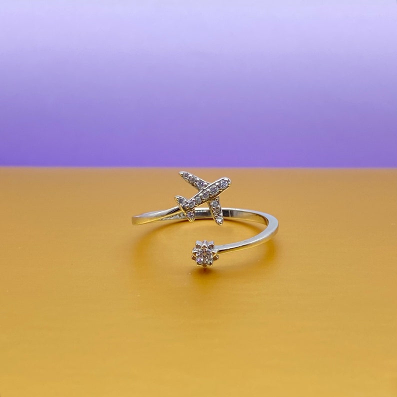Puede incluir: Anillo plateado con un peque&ntilde;o dise&ntilde;o de avi&oacute;n adornado con piedras preciosas transparentes. El anillo abierto presenta una piedra preciosa en el extremo de la banda. El fondo es un degradado de oro y p&uacute;rpura.