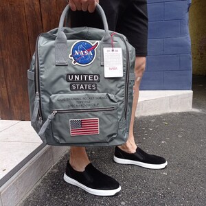 Puede incluir: Una mochila gris con parches de la NASA, un parche de la bandera estadounidense y el texto "UNITED STATES" y "CASE TRAINING, ROCKET SCIENCE TYPE RS2 SPEC No. 42-20074-P".