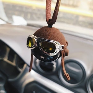 May include: A brown leather aviator-style car air freshener with a black and silver face and goggles. The air freshener is hanging from a rearview mirror.