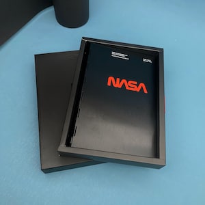 Könnte beinhalten: Eine schwarze Box mit silbernem Metallic-Finish und dem NASA-Logo in Rot. Die Box ist geschlossen und trägt auf der Vorderseite den Titel "National Aeronautics and Space Administration Graphic Standards Manual".