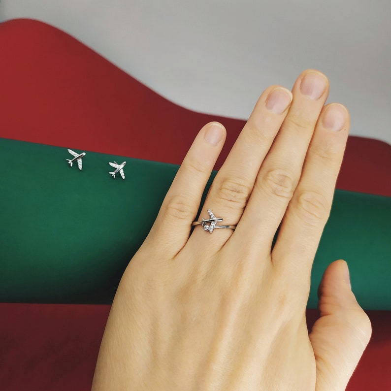 Op de afbeelding: Een zilveren vliegtuigring met een klein diamantje op de vleugel. De ring zit aan een vinger. De achtergrond is een groen en rood oppervlak.