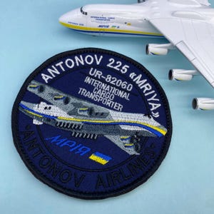 Może przedstawiać: Naszywka haftowana w kolorze niebieskim i czarnym przedstawiająca biały samolot z napisem "ANTONOV 225 «MRIYA»", "UR-82060", "INTERNATIONAL CARGO TRANSPORTER" i "МРІЯ" w białym i żółtym kolorze.