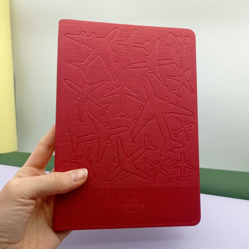 May include: Red notebook with a textured cover featuring an embossed design of multiple airplanes. The notebook has the text "AIR TRAILS" printed on the front cover.