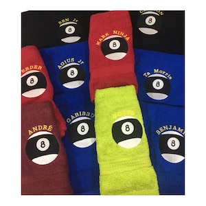 May include: A collection of eight personalised towels with embroidered names and a black and white 8-ball design. The towels are in various colours, including red, blue, green, and black.