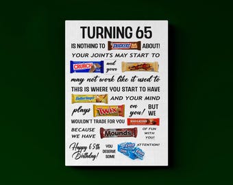 May include: A white rectangular sign with black text reading TURNING 65 IS NOTHING TO SNICKERS ABOUT, featuring various candy bar brand logos like Crunch, Butterfinger, Twix, 100 Grand, Mounds, and Extra gum integrated into the birthday message.