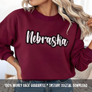 Op de afbeelding: Een bordeauxrode sweatshirt met het woord "Nebraska" in het wit, omlijnd met zwart, over de borst. De tekst heeft een distressed, vintage uitstraling. De sweatshirt is effen van kleur en heeft lange mouwen.