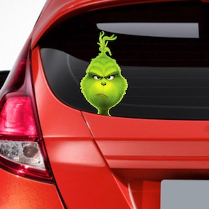 Könnte beinhalten: Ein grüner Grinch-Charakter-Aufkleber mit einem mürrischen Gesichtsausdruck, angebracht an der Heckscheibe eines roten Autos.