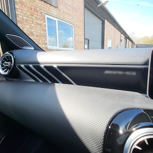 Puede incluir: Interior del salpicadero de un coche negro con detalles plateados. El salpicadero presenta una textura perforada y rayas plateadas. Se ve una salida de aire circular con ribete plateado. También está presente el logotipo de AMG.