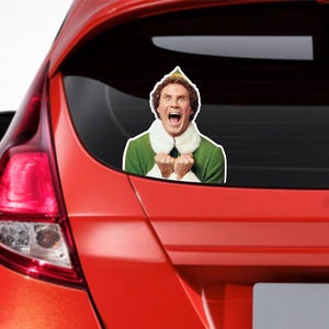 Op de afbeelding: Een rode auto met een Buddy the Elf-sticker op de achterruit. De sticker toont Buddy's gezicht met een verraste uitdrukking en zijn handen in de lucht.