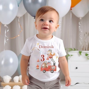 Könnte beinhalten: Ein weißes T-Shirt mit einem roten Feuerwehrauto und einem Fuchs darauf. Auf dem Shirt steht "Dominik 3". Der Junge trägt graue Shorts.