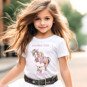 Op de afbeelding: Een jong meisje draagt een wit T-shirt met een roze eenhoorn en de tekst "Schulkind 2023" en de naam "Amelie". Ze draagt een witte tule rok en bruine cowboylaarzen.
