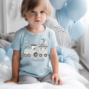 Könnte beinhalten: Ein hellblaues T-Shirt mit einem braunen Zugdesign mit Tieren und der Zahl 2. Das Shirt hat den Namen Dominik untenaufgedruckt.