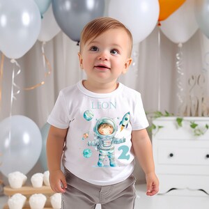 Könnte beinhalten: Ein Kind trägt ein weißes T-Shirt mit einem Weltraum-Motiv, das einen Astronauten, Planeten und eine Rakete zeigt. Auf dem Shirt stehen der Name "LEON" und die Zahl "2". Das Kind trägt graue Shorts und ist von Ballons umgeben.