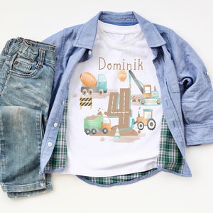 Könnte beinhalten: Ein weißes T-Shirt mit einem Bau-Motiv, das eine Nummer vier, Baufahrzeuge und den Namen Dominik zeigt. Das Shirt liegt auf einer weißen Oberfläche mit einer blauen Jeans und einem blau-grünen karierten Hemd.