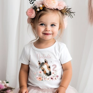 Könnte beinhalten: Ein weißes T-Shirt mit einem rosafarbenen Blumendesign und einer Illustration eines braunen und weißen Pferdes. Das Shirt trägt die Aufschrift "Natalie" und "2" in rosa Schrift.