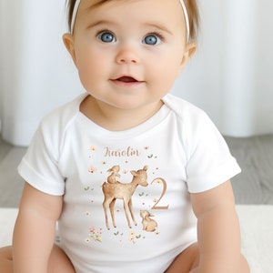 Op de afbeelding: Witte babybody met een illustratie van een hert en een konijn, de naam "Karolin" en het nummer "2". Het ontwerp bevat bloemaccenten in zachte kleuren. De baby draagt een witte haarband met een strik.
