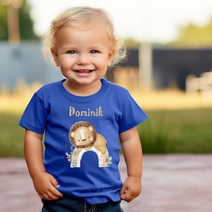 Könnte beinhalten: Ein königsblaues T-Shirt mit einer niedlichen Löwenillustration und dem Namen "Dominik" auf der Vorderseite.