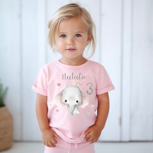 Könnte beinhalten: Ein rosa T-Shirt mit einem niedlichen Cartoon-Elefanten, der eine Krone trägt und die Zahl 3. Das Shirt trägt vorne den Schriftzug "Natalie".