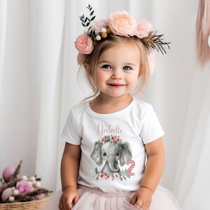 Könnte beinhalten: Ein weißes T-Shirt mit einem rosa Elefanten-Motiv und dem Namen "Natalie" darauf gedruckt. Der Elefant trägt eine rosa Blumenkrone und die Zahl "2" ist daneben gedruckt. Das Shirt wird von einem jungen Mädchen mit einer rosa Blumenkrone getragen.
