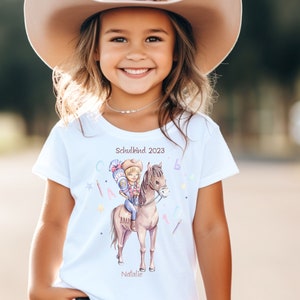 Könnte beinhalten: Weißes T-Shirt mit einer bunten Illustration eines Mädchens, das auf einem Pferd reitet. Der Text "Schulkind 2023" und "Natalie" sind auf das Shirt gedruckt.