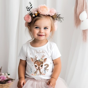 Könnte beinhalten: Ein weißes T-Shirt mit einer niedlichen Hirsch-Illustration und dem Text "Natalie" und der Zahl "2" in goldenen Glitzer. Das Shirt wird von einem jungen Mädchen mit einer Blumenkrone getragen.