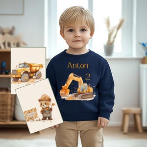Könnte beinhalten: Ein marineblaues Sweatshirt mit einem gelben Bagger und dem Namen Anton sowie der Zahl 2 auf der Vorderseite. Das Sweatshirt wird von einem kleinen Kind gehalten.