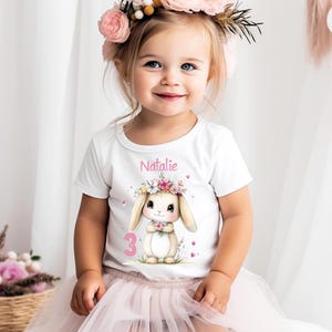 Könnte beinhalten: Ein junges Kind trägt ein weißes T-Shirt mit einem Hasen-Motiv und dem Namen "Natalie". Der Hase hat einen Blumenkranz und hält Blumen. Die Zahl "3" befindet sich unter dem Hasen. Das Kind trägt einen rosa Tüllrock und einen Blumenkranz.