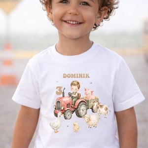 Könnte beinhalten: Weißes T-Shirt mit einer Aquarellillustration eines roten Traktors, der von einem Jungen gefahren wird, mit einem Schwein, einer Kuh und einem Schaf. Der Name "Dominik" und die Zahl "3" sind über dem Traktor aufgedruckt.