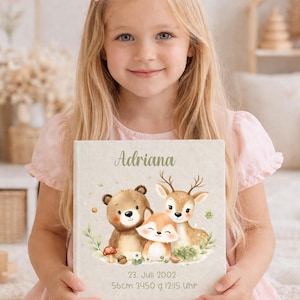 Könnte beinhalten: Ein hellbeige Baby-Erinnerungsbuch, das von einem jungen Mädchen gehalten wird. Das Cover zeigt eine Aquarellillustration eines Bären, eines Fuchses und eines Hirsches. Der Name "Adriana" steht in grüner Schrift über der Illustration. Das Buch enthält auch Datum, Uhrzeit und Maße.