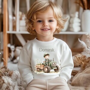 Könnte beinhalten: Weißer Pullover mit einem grünen Traktor und einem Jungen, der ihn fährt. Der Text "Dominik" ist auf der Vorderseite des Pullovers gedruckt.
