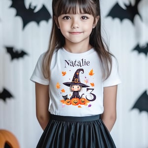 Könnte beinhalten: Ein weißes T-Shirt mit dem Aufdruck "Natalie", das ein Halloween-Motiv zeigt. Das Motiv zeigt eine Hexe, Kürbisse, eine schwarze Katze und die Zahl "3". Das Mädchen trägt einen schwarzen Rock.