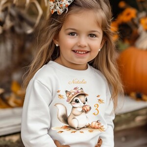 Könnte beinhalten: Ein junges Mädchen trägt ein weißes Sweatshirt mit einem Eichhörnchen-Motiv, das einen Kürbis hält. Der Aufdruck enthält den Namen "Natalie" und die Zahl "3". Das Mädchen trägt auch eine Blumen-Schleife und Ohrringe. Der Hintergrund zeigt Kürbisse und Herbstlaub.