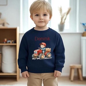 Könnte beinhalten: Ein marineblauer Pullover mit einem roten Traktor und einem Jungen, der ihn fährt. Der Text "Dominik" ist auf der Vorderseite des Pullovers aufgedruckt.