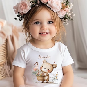 Könnte beinhalten: Ein weißes T-Shirt mit einem braunen Bären, der einen Korb mit Blumen hält. Das Shirt ist mit dem Namen "Natalie" und der Zahl "2" bedruckt. Das Mädchen trägt eine Blumenkrone.
