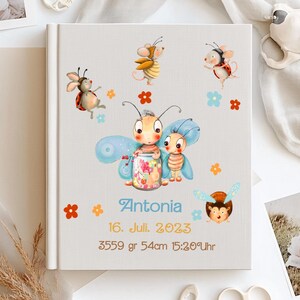 Peut inclure: Un album de souvenirs pour bébé de couleur claire, avec des illustrations d'insectes et de fleurs de dessins animés. La couverture présente le nom "Antonia" et la date "16. Juli. 2023", ainsi que les mesures de poids et de taille. Le livre est décoré d'accents colorés.