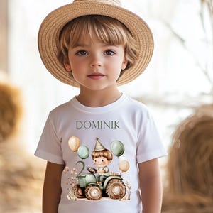 Könnte beinhalten: Weißes T-Shirt mit einem grünen Traktor und einem Jungen, der ihn fährt. Der Traktor hat die Nummer 2 an der Seite und ist von Ballons umgeben. Der Text "DOMINIK" ist auf das Shirt gedruckt.