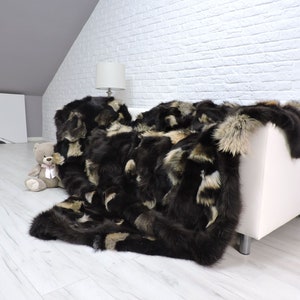 May include: A black and brown faux fur throw blanket with a shaggy texture. The blanket is draped over a white couch and a stuffed animal is on the floor.