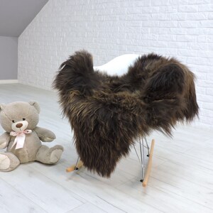 May include: A brown sheepskin rug draped over a white rocking chair. A tan teddy bear with a pink bow sits on the floor next to the chair. The room has white brick walls and light wood floors.