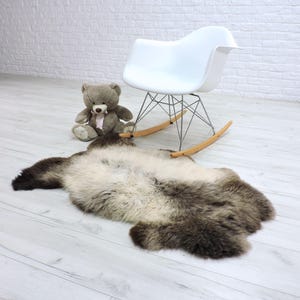 Op de afbeelding: Een schapenvacht tapijt met bruine en crèmekleurige tinten ligt op een lichte houten vloer. Achter het tapijt staat een witte schommelstoel met een metalen frame en houten schommels. Een grijze teddybeer zit in de buurt.