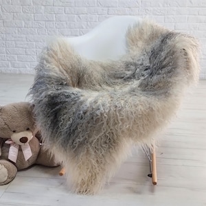 Op de afbeelding: Een langharig, krullend schapenvachtkleed met een verloop van crème, beige en antracietgrijze tinten. Het kleed is gedrapeerd over een witte stoel, naast een lichtbruine pluche teddybeer met een roze lint om de nek gestrikt.