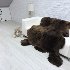 Pode incluir: Um tapete de pele de ovelha castanho escuro sobre um sofá branco. O tapete tem uma aparência espessa e texturizada. Um urso de pelúcia cinzento com um laço rosa está sentado no chão perto do sofá. Um retrato a preto e branco está pendurado na parede.