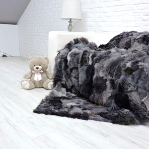 Peut inclure: Une luxueuse couverture en fausse fourrure gris foncé drapée sur un canapé blanc. La couverture présente un mélange de tons gris et noirs, créant un aspect texturé. Un ours en peluche avec un nœud rose est à proximité. La pièce a un mur de briques blanches.