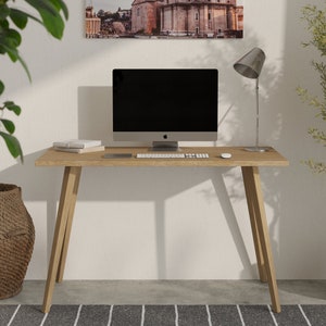 Könnte beinhalten: Ein heller Holzschreibtisch mit einem silbernen Laptop, einer Tastatur und einer Maus. Eine Lampe mit einem silbernen Schirm steht auf der linken Seite des Schreibtisches. Eine Topfpflanze steht auf der rechten Seite des Schreibtisches. Ein Weidenkorb steht auf dem Boden links vom Schreibtisch.
