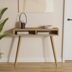 Może przedstawiać: Jasny drewniany stolik konsolowy z dwoma otwartymi półkami. Stolik ma minimalistyczny design z zwężającymi się nogami i jest ozdobiony czarnym świecznikiem, stosem książek i ozdobną misą.