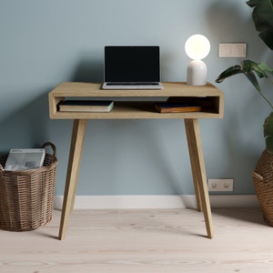 Könnte beinhalten: Ein heller Holzschreibtisch mit einem Laptop, zwei Büchern und einer weißen Globuslampe. Der Schreibtisch hat ein Regal darunter und steht auf einem hellen Holzboden. Auf dem Boden stehen zwei Weidenkörbe, einer mit einer Zeitschrift darin.