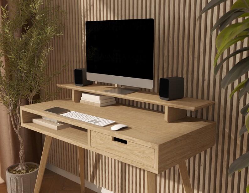 Puede incluir: Un escritorio de madera clara con un caj&oacute;n y un estante para un monitor de ordenador. El escritorio tiene un dise&ntilde;o moderno con patas inclinadas y un aspecto simple y limpio. El escritorio est&aacute; colocado contra una pared de listones de madera con una planta en maceta en el lado izquierdo.