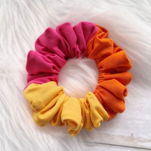 Pode incluir: Um scrunchie de cabelo colorido feito de tecido em tons de rosa, laranja e amarelo. O scrunchie &eacute; reunido em forma de c&iacute;rculo.