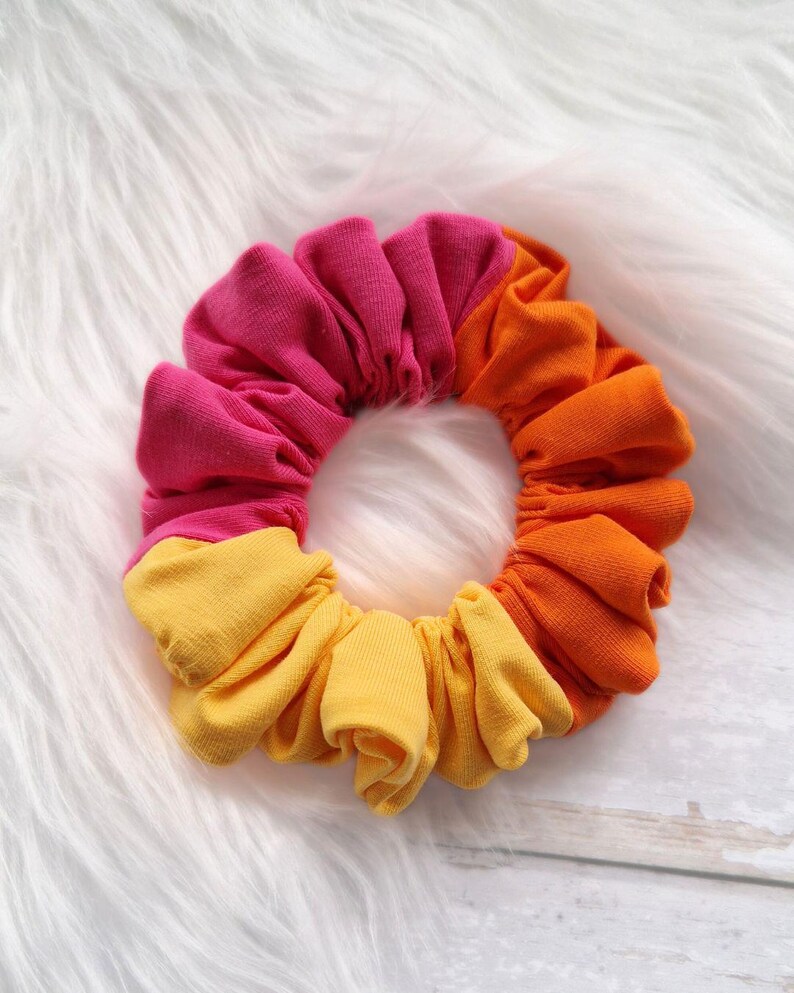 Pode incluir: Um scrunchie de cabelo colorido feito de tecido em tons de rosa, laranja e amarelo. O scrunchie &eacute; reunido em forma de c&iacute;rculo.