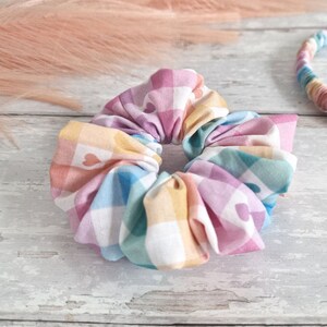 May include: A colorful scrunchie with a checkered pattern and small white hearts. The scrunchie is made of fabric and is in a pastel color scheme.
